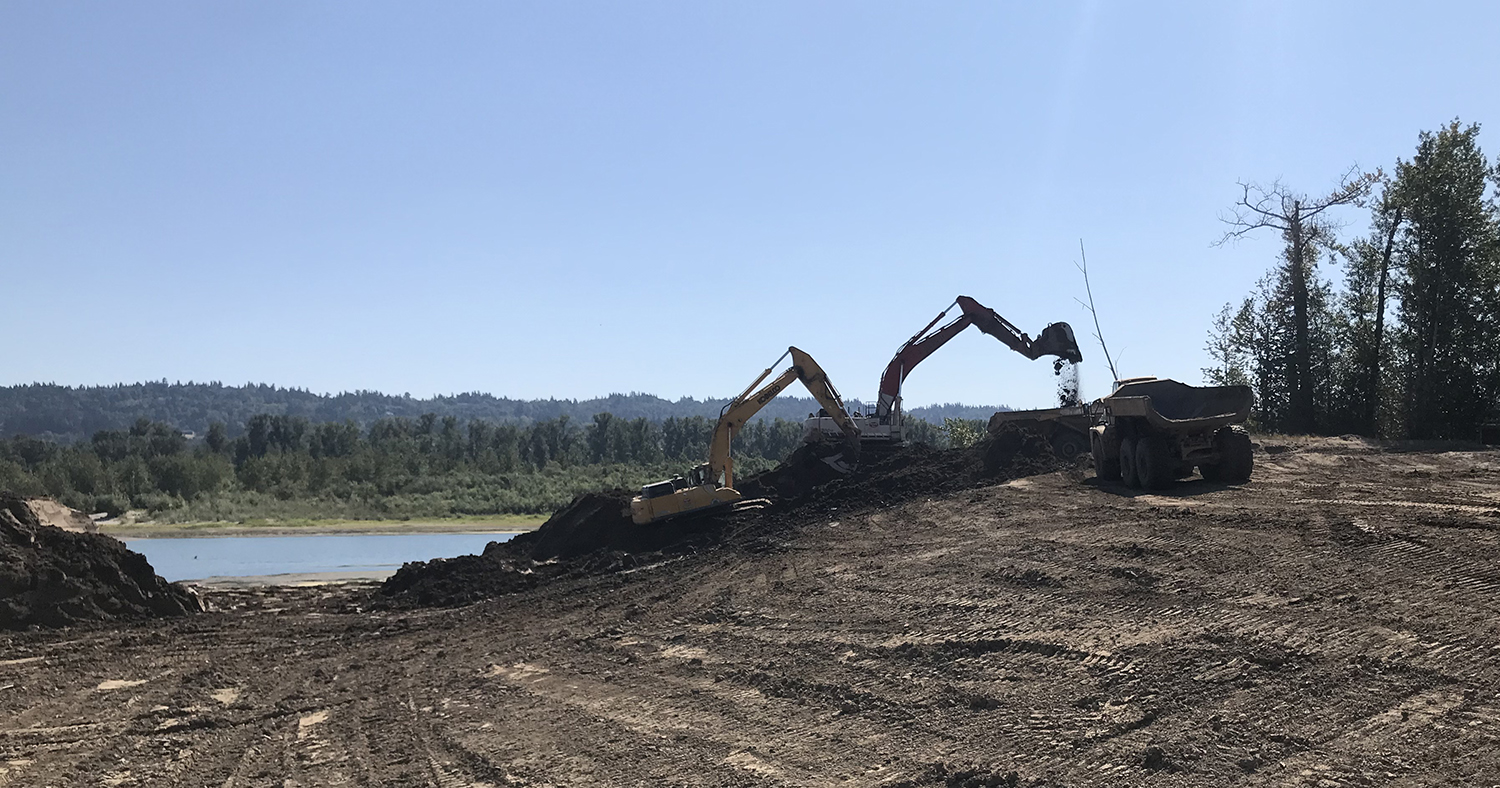 An excavator digs through a massive levee. The Columbia River is visible through the breach.