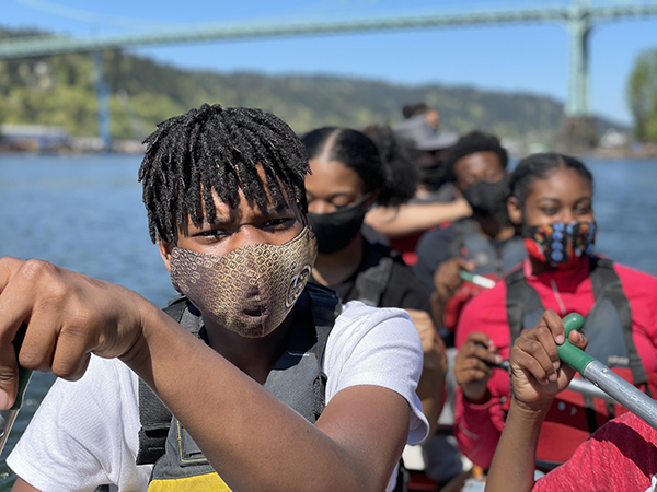 a young man in a mask looks into the camera while he paddles a big canoe with others.