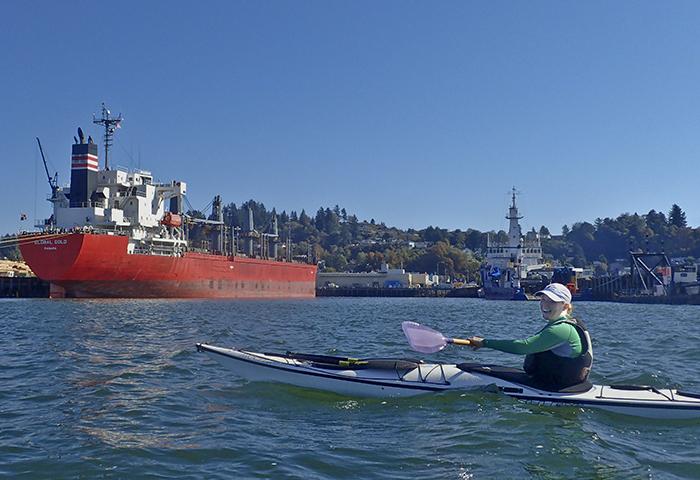 woman kayaks along the Astoria waterfront and a large cargo ship named Global Gold