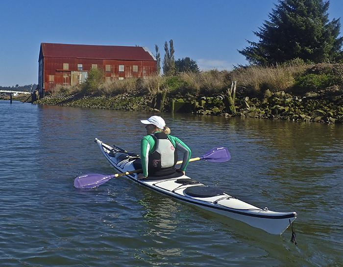 woman kayaks Youngs Bay toward a large red building