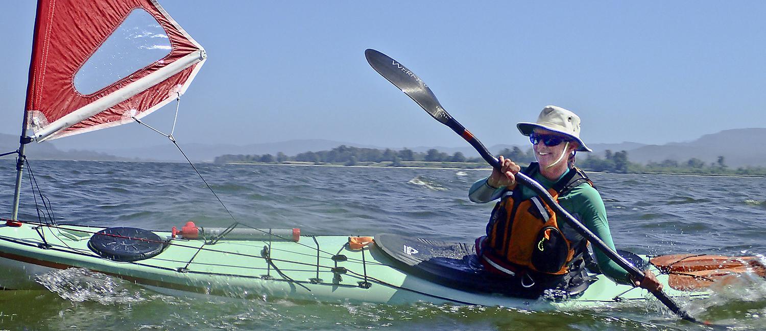 Ginni smiling, paddling her kayak with a sail on the front.