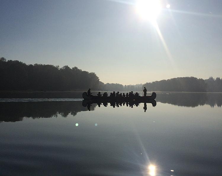 silhouette of a big canoe carrying about 14 people. The water is smooth and the sun is reflecting off it.