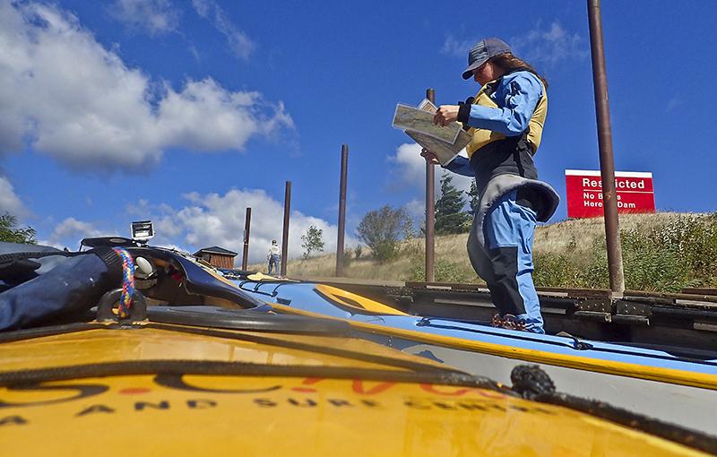 woman stands by a kayak wearing a pfd and spray skirt and looking at a printed chart