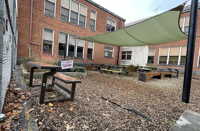 outdoor courtyard with wood picnic tables and a raingarden, covered by a shade sail