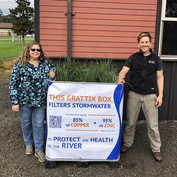 Two people pose with a Grattix box, which reads "This Grattix Box filteres stormwater. It removes up to 85% + 95% of zinc from runoff to protect the health of the river"