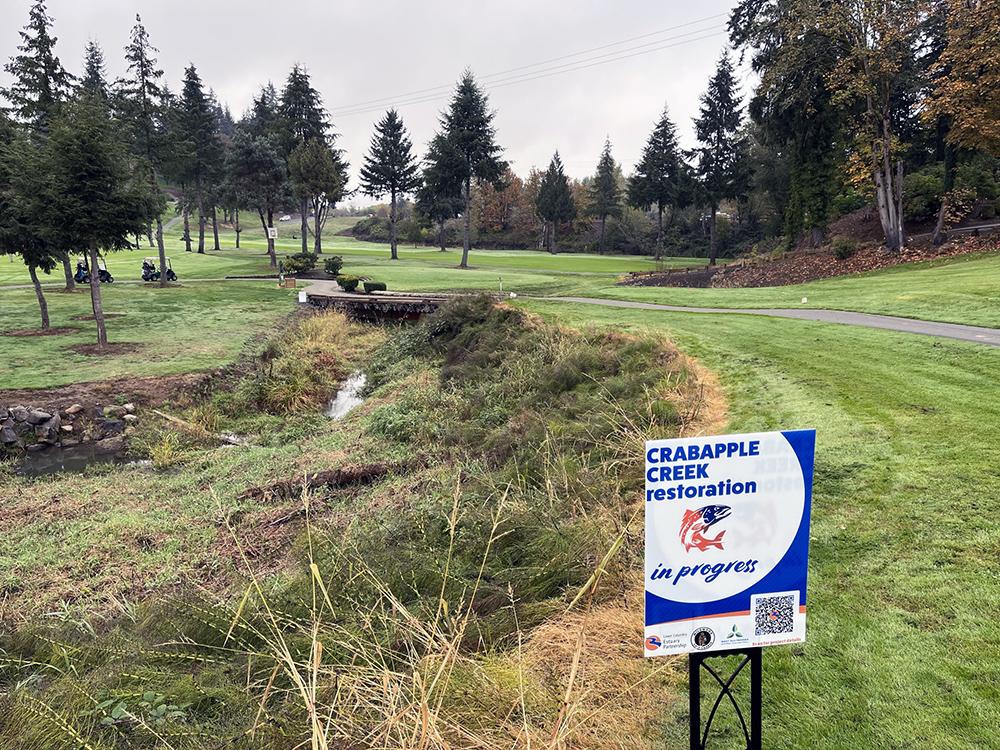 a yard sign for the restoration project by the creek bed, with golf carts in the distance