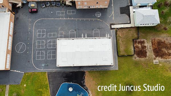 aerial view of the schoolyard shows a large asphalt parking lot and play area, including a covered area, and adjacent grassy areas and buildings