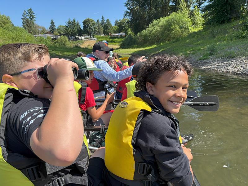 two kids in the back of a big canoe, one using binoculars and the other smiling