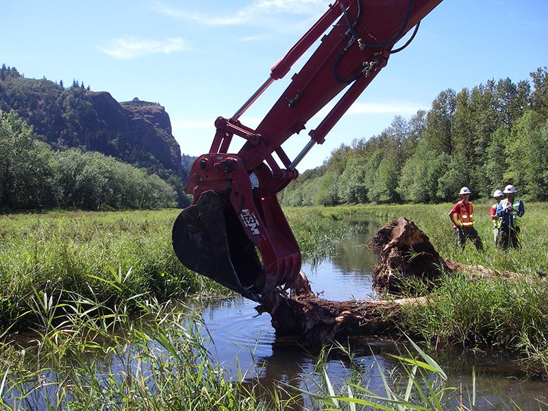 a material handler truck arm places down a large log into a lake with three people looking on