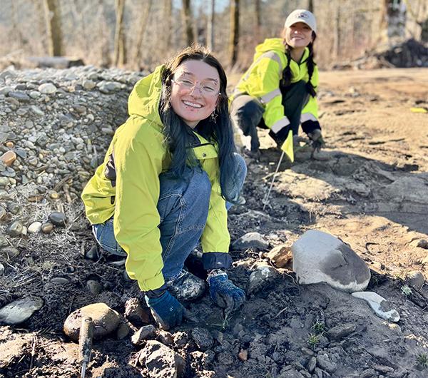 two volunteers pause to smile while crouching down to plant 