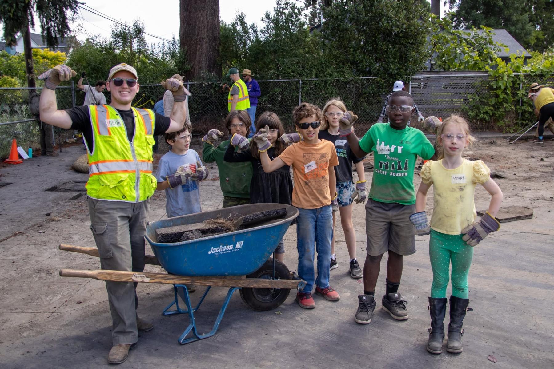an adult and several kids stand around a wheelbarrow full of asphalt bits and pose with a bicep curl 