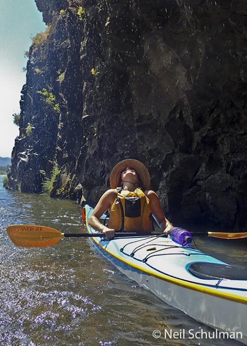 woman in a kayak has her head back, enjoying drips from a small waterfall