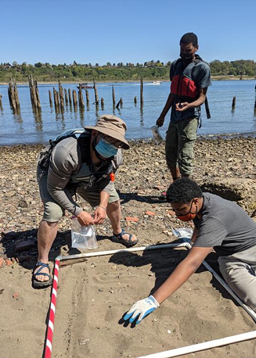 a boy searches within a sandy plot for microdebris. An instructor leans over, and another boy waits to show an item.