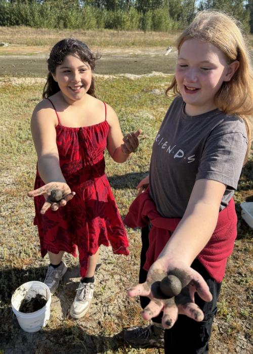 two students hold muddy wapato seed balls